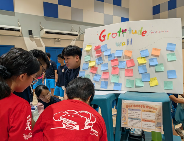 Hai Sing Catholic School students in red and blue PE shirts writing on colorful sticky notes and placing them on a decorated "Gratitude Wall" board during HAI Spirits Week.